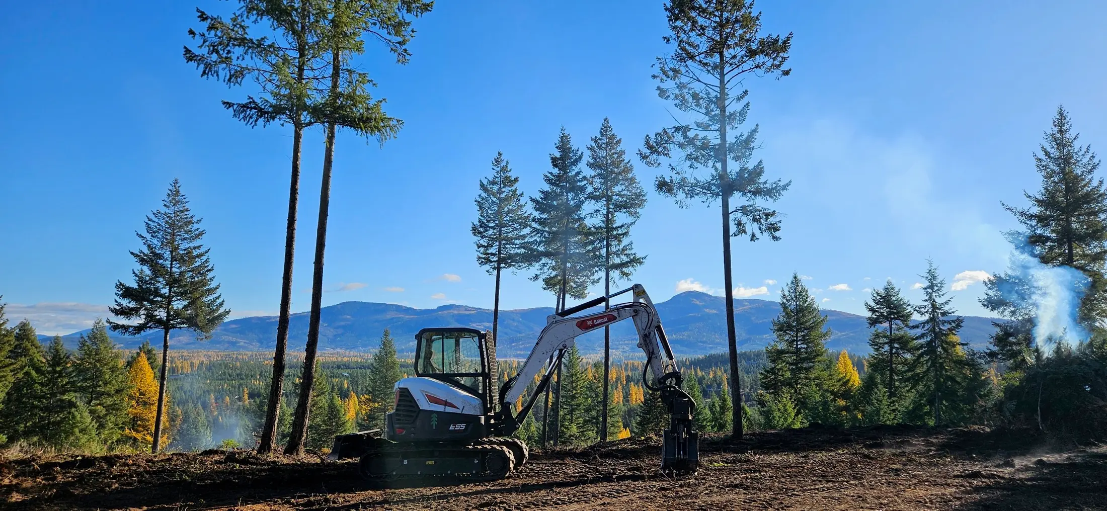 Freshly thinned and cleared ridge above the valley, excavator on site, Selkirk foothills behind